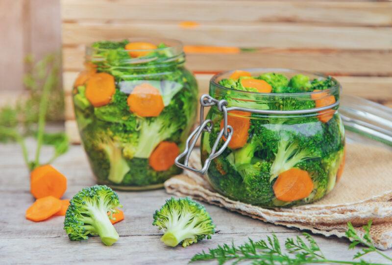 a couple of jars of fermented broccoli florets with carrots slices, with loose broccoli florets, carrot slices, and herbs around it