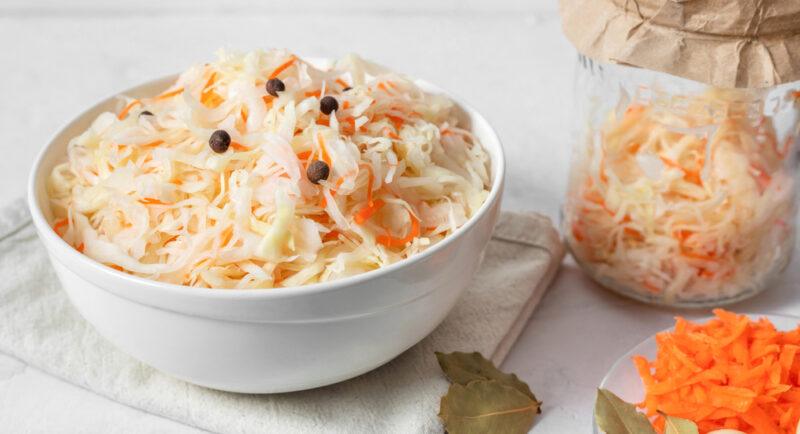 on a white surface is a white ceramic bowl full of fermented cabbage resting on top of a beige table napkin with a bay leaf and a white saucer with shredded carrots beside it, at the back is a jar with fermented cabbage as well