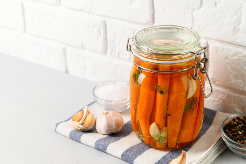 a jar of fermented whole carrots with deal, garlic cloves, and dill, resting on top of a striped white and blue table napkin with a couple of garlic heads, a small bowl of rock salt, and a little bowl of black whole peppercorns beside it