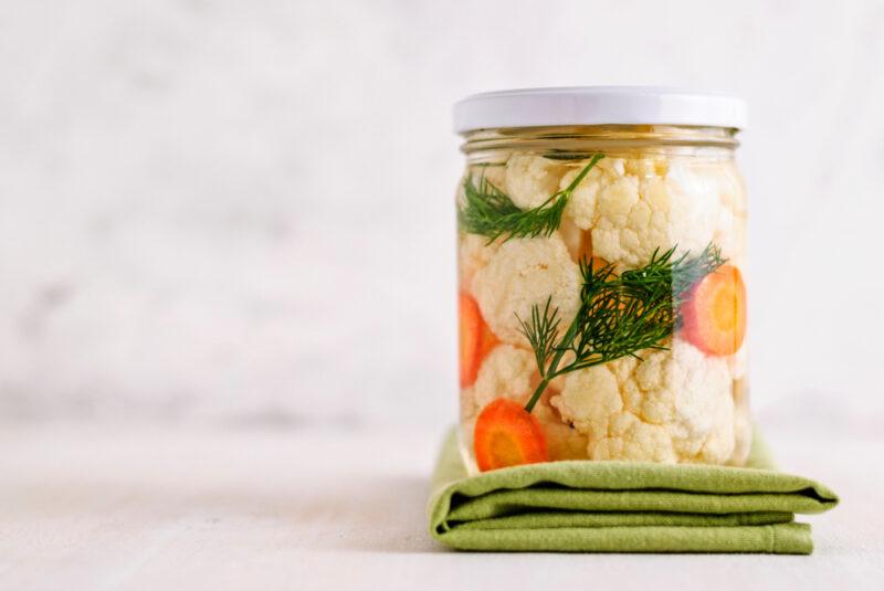 on a white surface is a jar of cauliflower floret with carrot slices and fresh dill, resting on top of a green table napkin