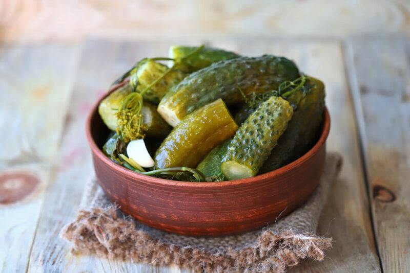 on rustic looking wooden surface is a dark brown wooden bowl full of fermented cucumbers with a clove of garlic and sprigs of dill and dill flowers, resting on a burlap