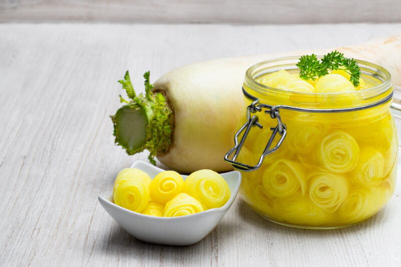 on a white wooden surface is a jar of fermented daikon with a small white dish with fermented daikon, too. At the back is a whole fresh daikon