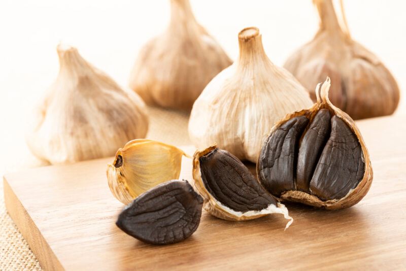 on a wooden chopping board are a few fermented garlic heads, with one partly cracked showing off its black color