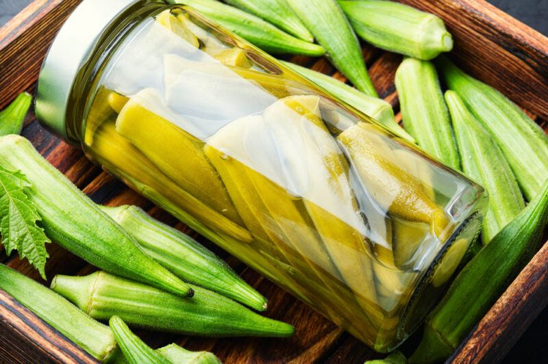 a square wooden tray with a jar of fermented okra on its side with fresh okra around it