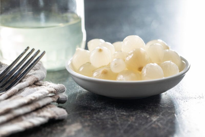 on a marble surface is a white saucer with fermented white onions with a fork resting on top of a napkin, at the back is a jar with brine in it