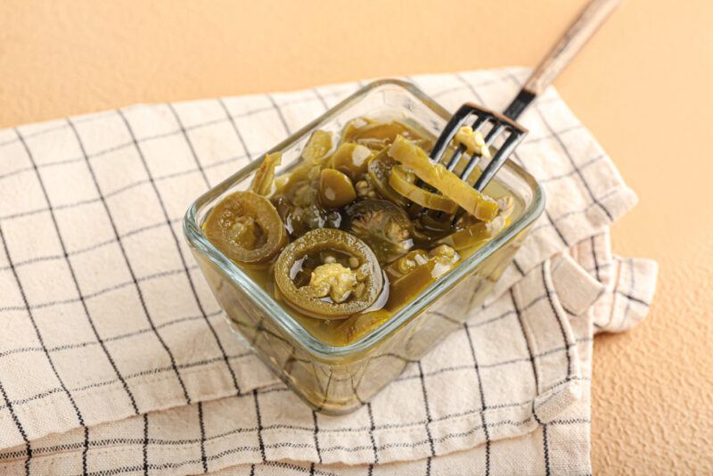 on beige concrete surface is a transparent glass square dish full of fermented pepper, jalapeno, with a fork in it, resting on top of a white table napkin with blue stripes