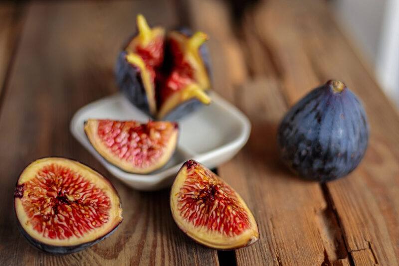 on a rustic looking wooden surface is a small white square dish with cut figs, beside it is a whole fig, and figs wedges at the foreground