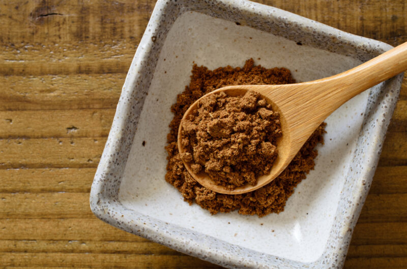 on a wooden surface is a white stone square dish with gray rim, holding a small heap of five spice with a wooden spoon on top of it also with five spice