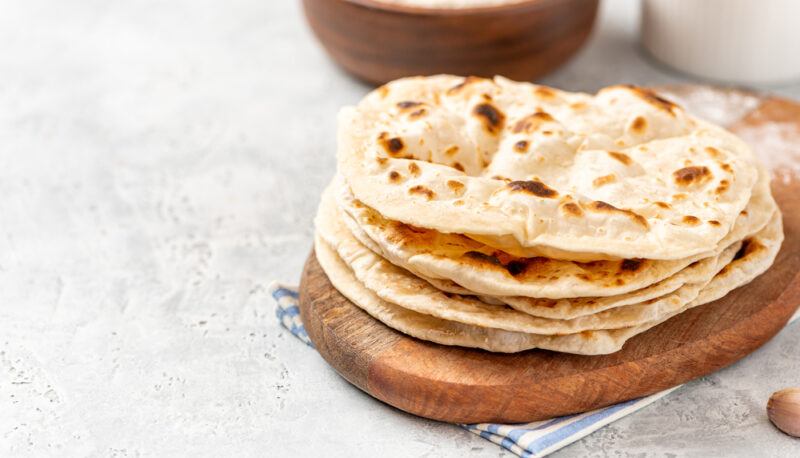 on a marble surface is a wooden chopping board with a pile of flatbread, behind is a partial image of brown bowl