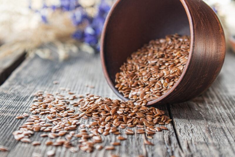 on a rustic wooden table is a brown bowl spilled over with its flaxseeds contents pouring out