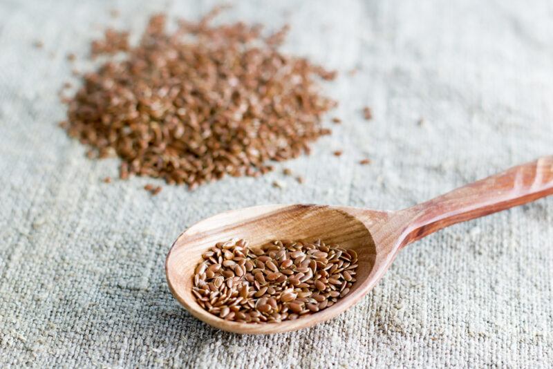 on a light grey textile is a wooden spoon with flaxseed, at the back is a small mound of flaxseed