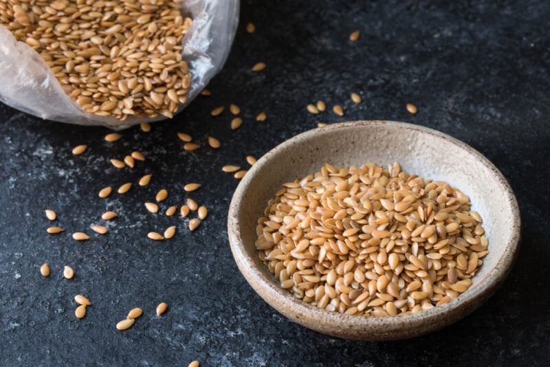 on a black concrete surface is a bowl of flaxseeds with a plastic bag of flaxseeds behind it and loose flaxseeds around them