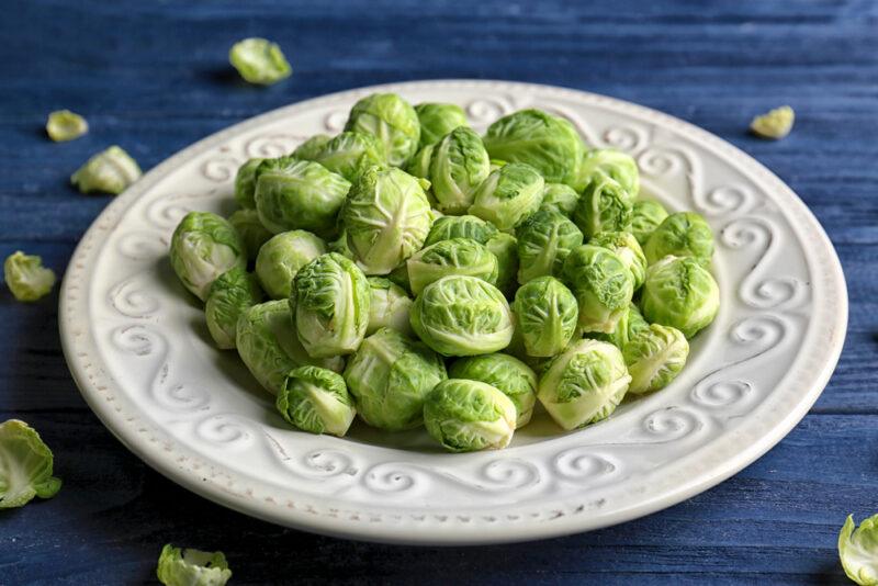 on a blue wooden surface is a white plate full of fresh Brussels sprouts with loose Brussels sprouts around it