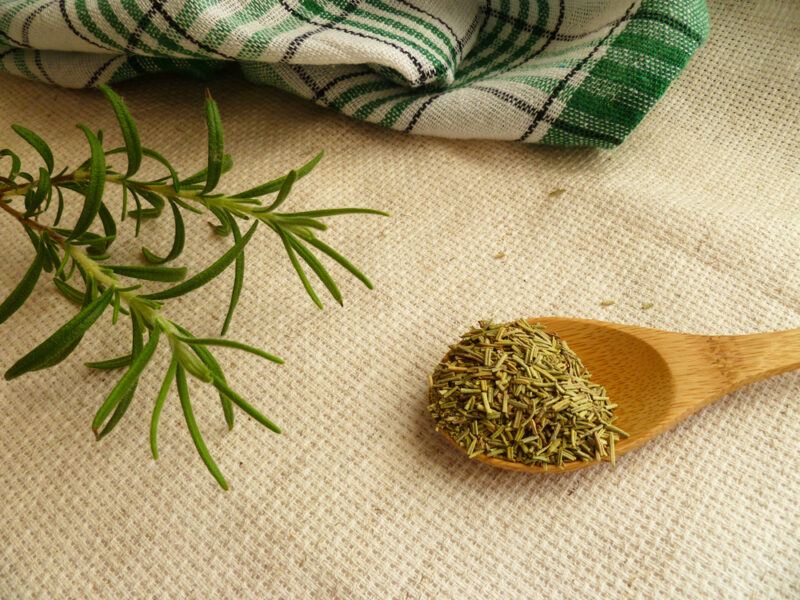 on a brown place mat is wooden spoon full of dried rosemary, beside it are fresh rosemary with a white and green table napkin at the back