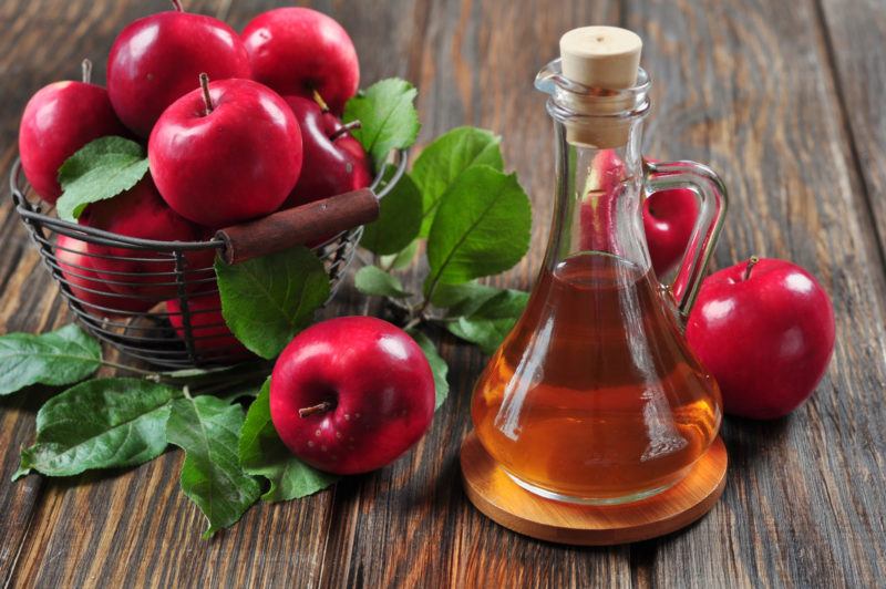 fresh red apples in a wire basket and a glass container filled with apple cider on a wooden table