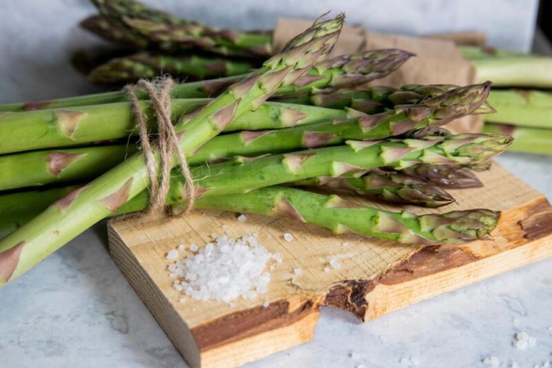 on a white surface is a wooden chopping board with a small mound of rock salt and a couple of bunch of fresh asparagus