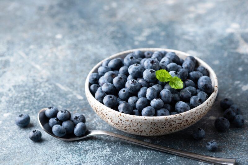 on a concrete surface is a clay bowl full of fresh berries with a sprig of mint on top, a silver spoon in front of it with blueberries can be seen as well as loose blueberries around it