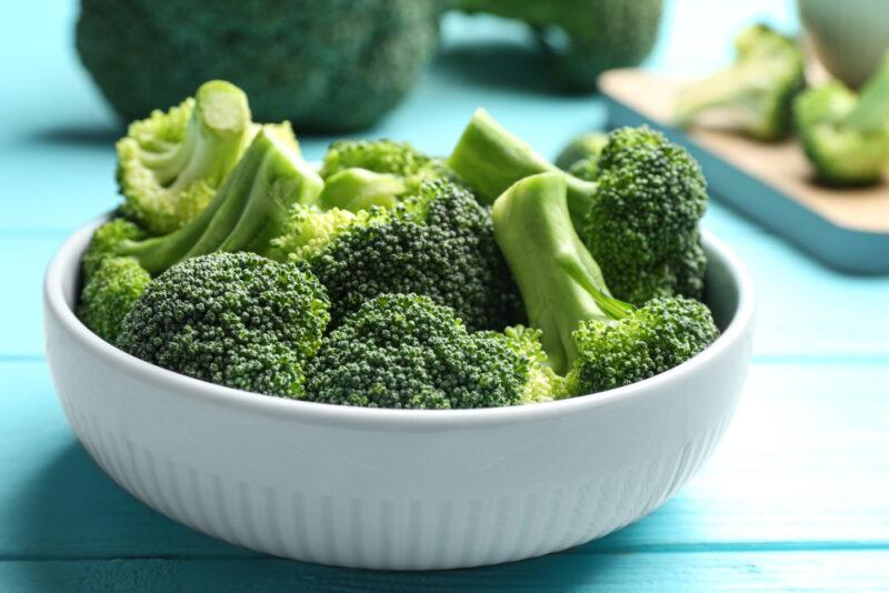 on a blue wooden surface is a white bowl full of broccoli florets, at the back is a partial image of a wooden chopping board with broccoli florets on it, beside it is a partial image of a head of broccoli