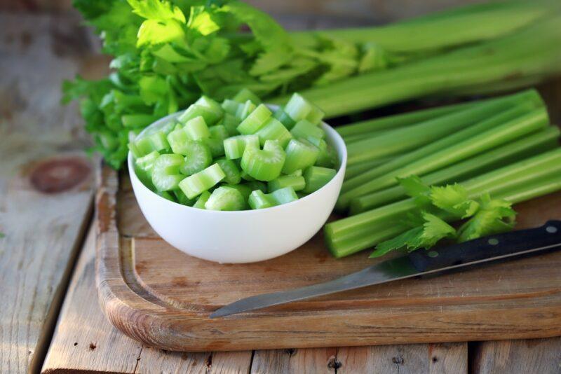 on an aged wooden surface is a wooden chopping board with a white bowl of chopped celery and celery sticks beside it