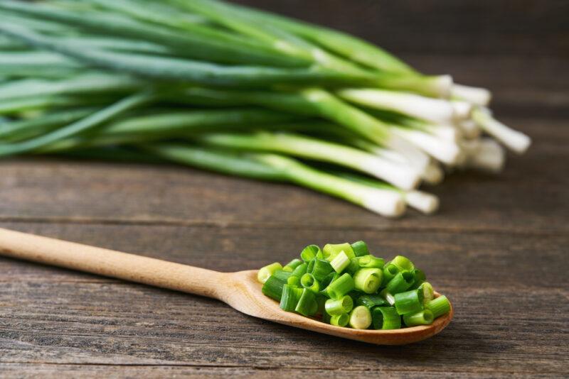 on a rustic looking wooden surface is a wooden spoon full of minced fresh chives, at the back is a small pile of fresh chives as well