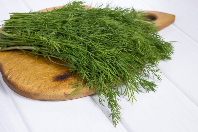 on a white wooden surface is a brown wooden chopping board with fresh dill on it