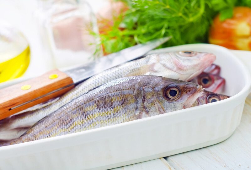 closeup image of a white ceramic dish with fresh whole fish, a knife and fresh herbs can be seen at the background 