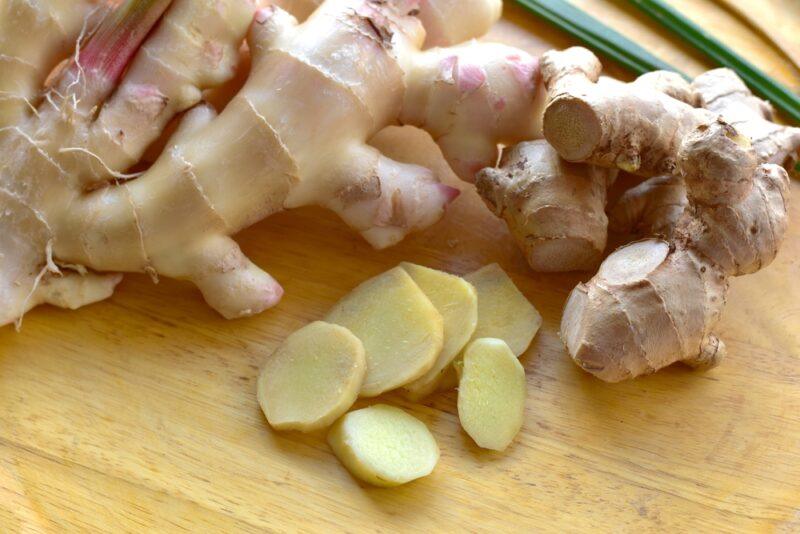 on a wooden surface is a few slices of fresh ginger and whole ginger rhizomes behind it