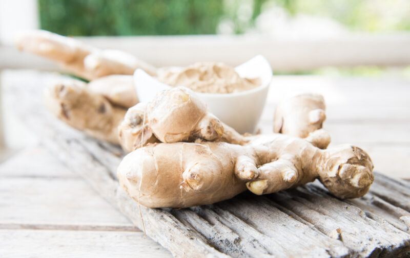 on a wooden surface is a rustic wooden plank with fresh ginger roots on it and white dish full of ground ginger at the back