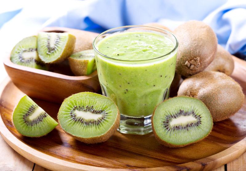 Kiwi fruits, some sliced in halves, some whole, plus a glass of fresh kiwi juice, resting on a wooden platter with light blue cloth at the back