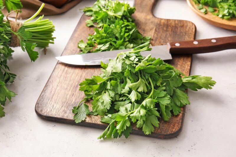 on a marble surface is a wooden chopping board with chopped fresh parsley and a knife on it, around it are partial image of a bunch of fresh parsley and a dish with chopped parsley