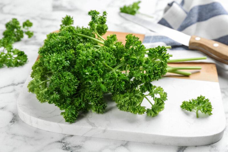 on a marble surface is a marble chopping board with fresh parsley on it, with white and blue table napkin, knife, and loose fresh parsley at the back