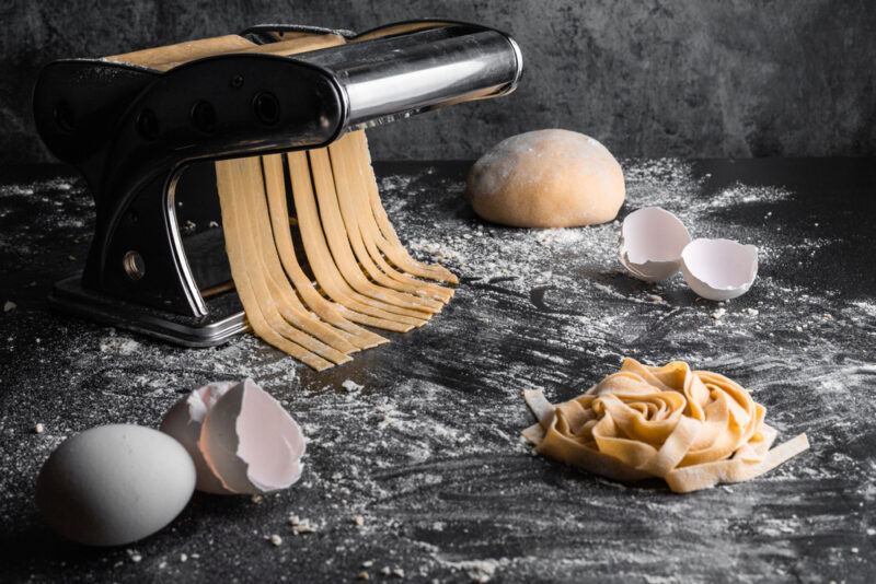 on a black surface are fresh pasta coming out of a pasta maker, fresh dough can be seen at the back, a mound of fresh pasta and one whole egg, eggshells, and loose flour around