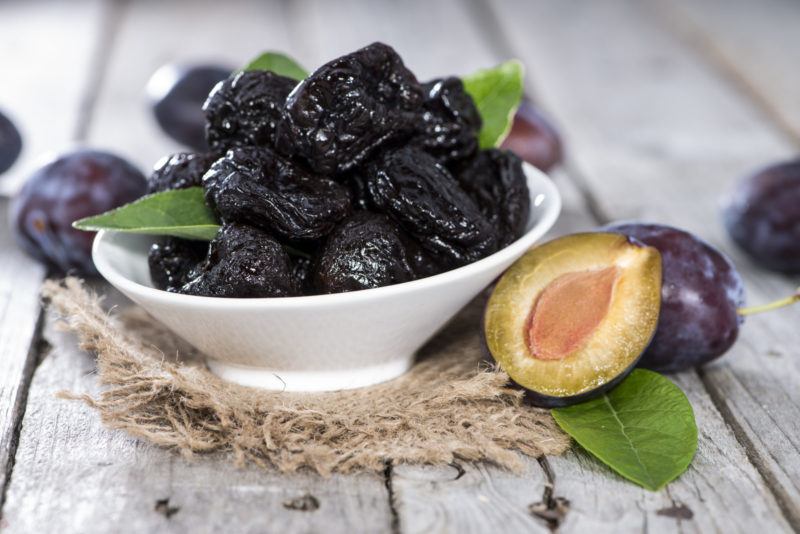 a shallow white bowl full of prunes plus a fresh cut plum resting on a piece of burlap sack on an aged-wooden surface