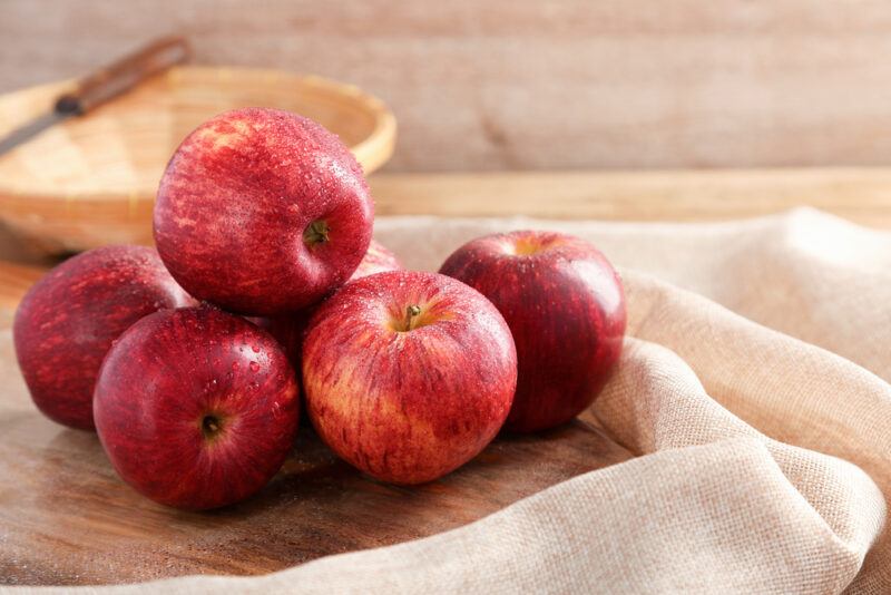 on a wooden surface is  a beige cloth  beside a wooden chopping board with a pile of fresh red apples, at the back is a bowl with a knife on it