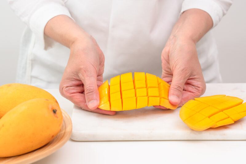 woman's hands holding a sliced mango over a white chopping board with sliced mango, beside it are whole mangoes