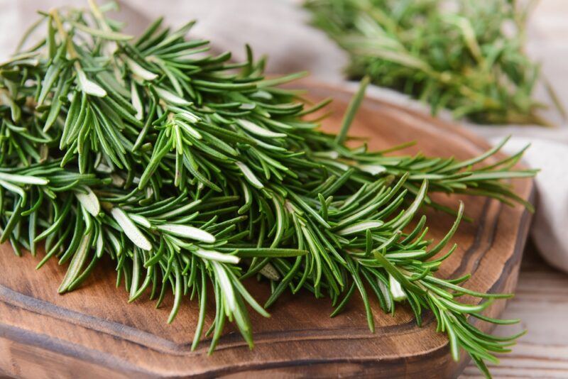 wooden surface is a closeup image of a wooden chopping board with a bunch of fresh rosemary, at the back is a sprig of fresh rosemary as well