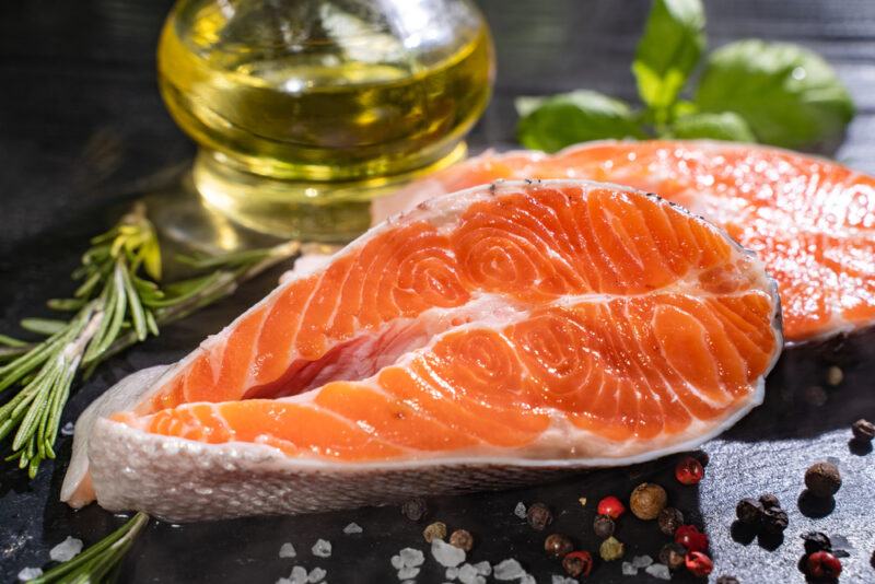 on dark surface is a fresh slice of salmon with loose herbs, rock salt, and black and pink peppercorns around it, at the back is also a bottle of olive oil