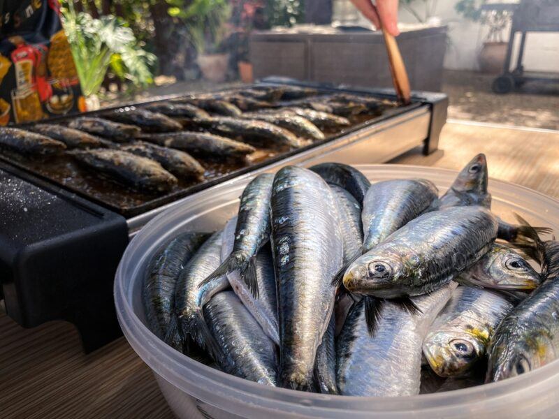 a plastic round bowl full of fresh sardines which is a kind of fatty fish, behind is a grill with fresh sardines being cooked