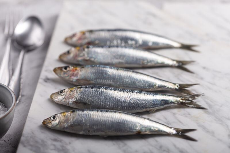 fresh sardines neatly laid on a marbled board, with a silver spoon and fork and partial image of a small bowl