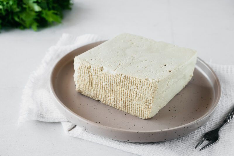 on a white surface is a tan colored dish with a block of fresh tofu resting on a white table napkin with a black two-prong fork, at the back is a bunch of herbs