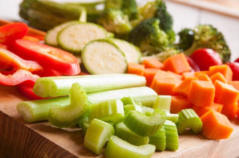 a closeup image of a chopping board with chopped vegetables, celery, carrots, red bell pepper, zucchini, and broccoli 