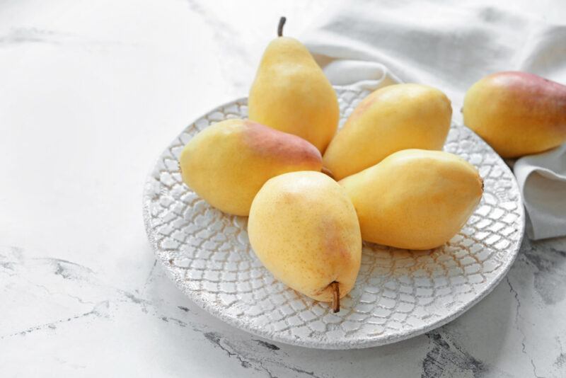 on a marbled surface is a white textured dish with five whole pears, with a white table napkin with one whole pear behind it.