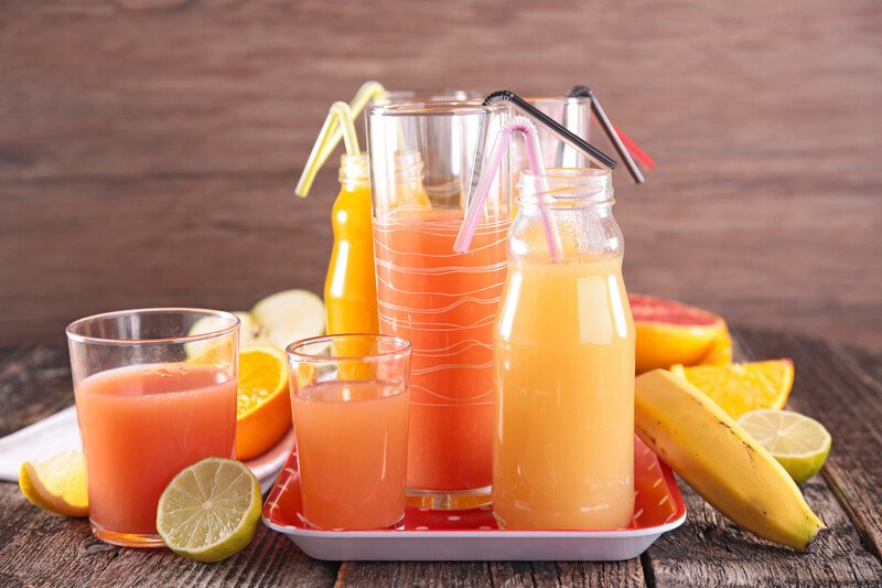 on a wooden surface and wooden background are different glasses with straws full of different fruit juices, with fresh fruits at the back