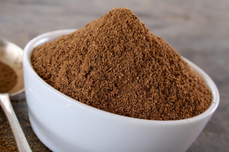 a closeup image of a white bowl full of garam masala with a metal spoon beside it