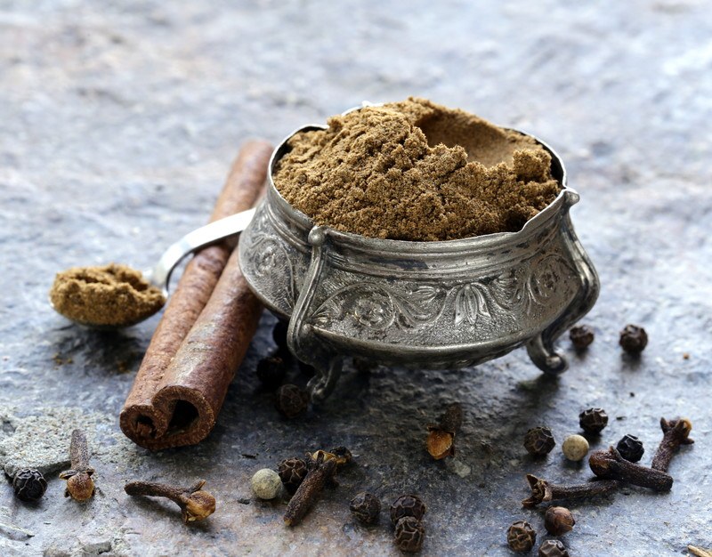 a unique metal container full of garam masala resting on a marble surface with cinnamon sticks, cloves, peppers, and a metal spoon full of garam masala