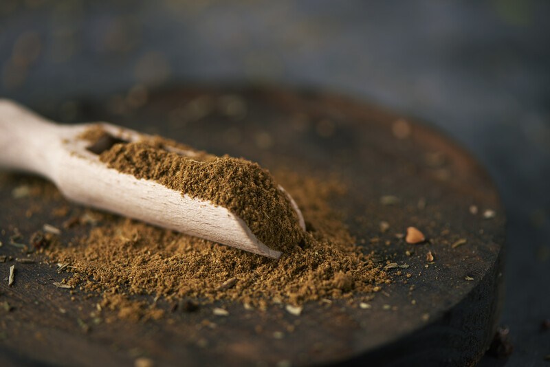 closeup image of a round wooden board with a wooden scoop full of garam masala with garam masala resting on a small mound of garam masala