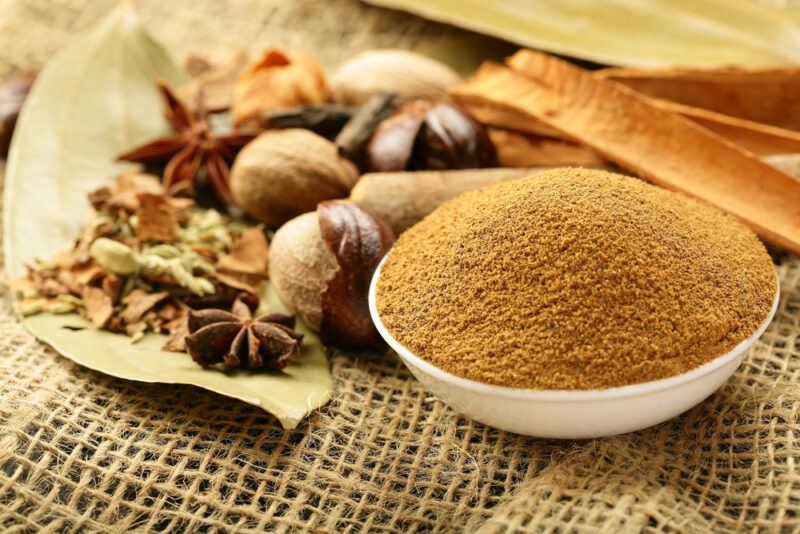 laid on a burlap is a white bowl full of garam masala and different whole spices behind it like cinnamon sticks, star anise, bay leaves, and more