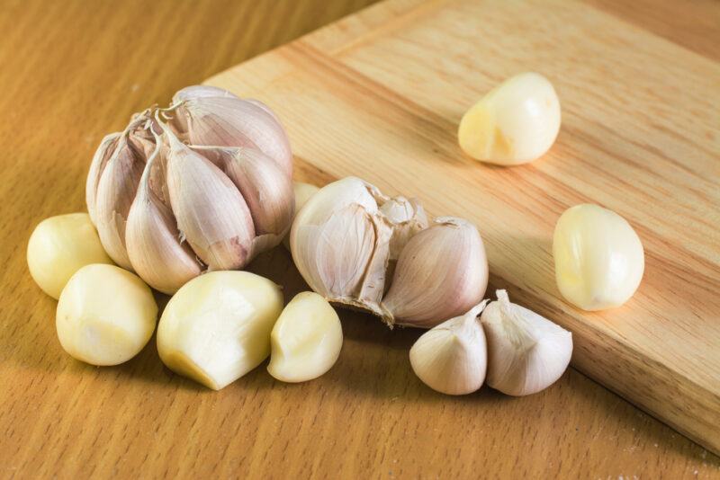 on a wooden surface is a couple of garlic head with loose garlic cloves, some are peeled, behind it is a wooden chopping board with a couple of peeled garlic cloves