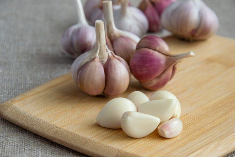 on a grey concrete surface is a wooden chopping board with peeled garlic cloves and garlic heads