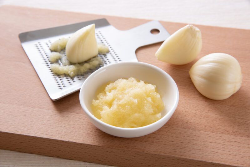 on a wooden chopping board with a small white dish with grated garlic, behind it are few garlic cloves and a metal grater